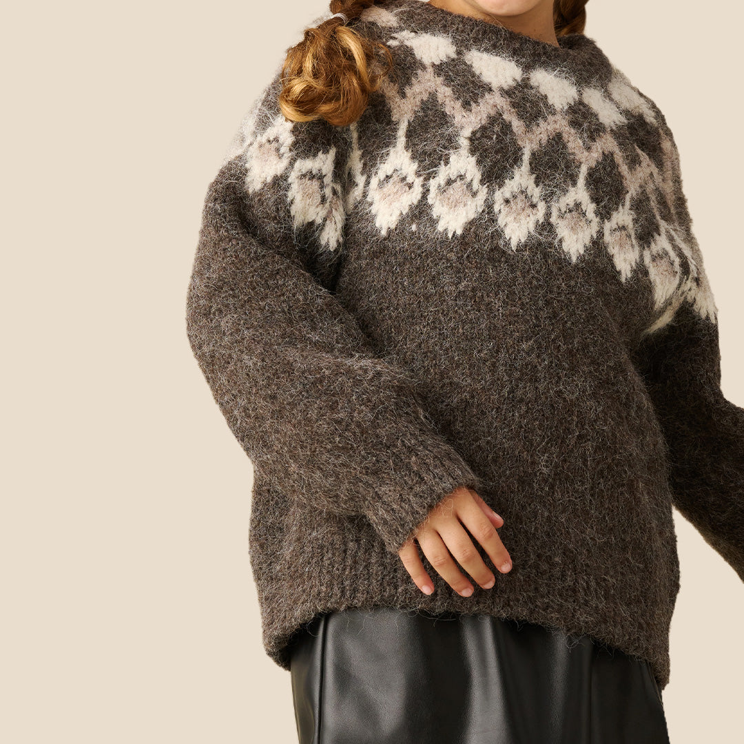 A young girl modeling a chunky knit crewneck sweater with a brown and cream argyle pattern, paired with a black faux-leather mini skirt, against a solid beige background.