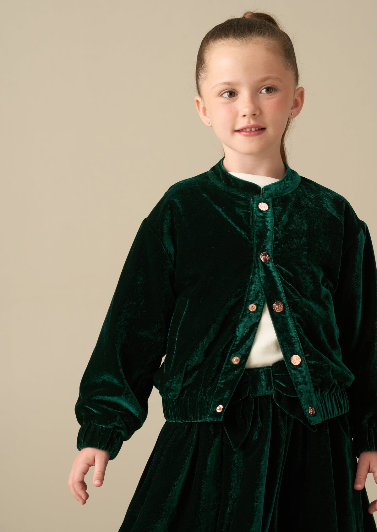A young girl with her hair in a ponytail wearing a dark green velvet jacket and matching skirt over a white top, photographed against a solid beige studio background.