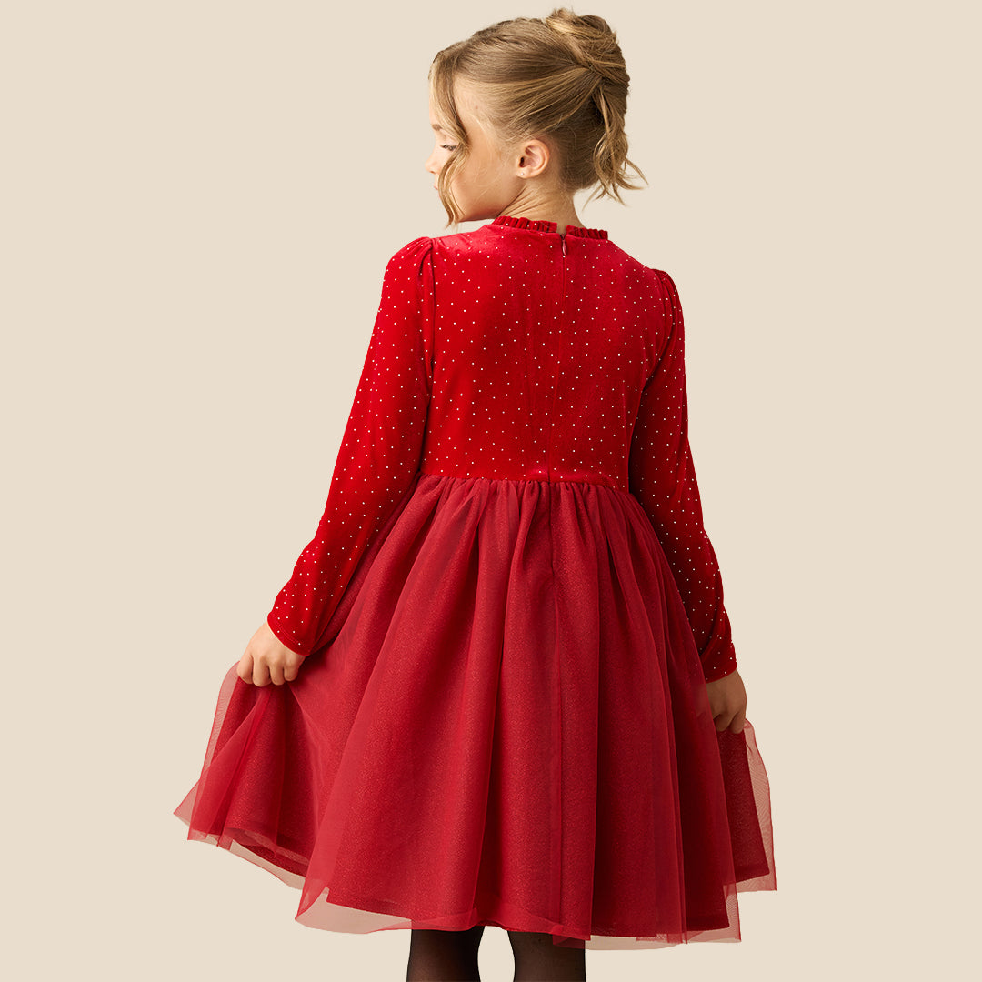 A young girl wearing a festive red dress with long sleeves, tiny gold polka dots, a matching red bow at the waist, and a tulle skirt, set against a solid beige background.