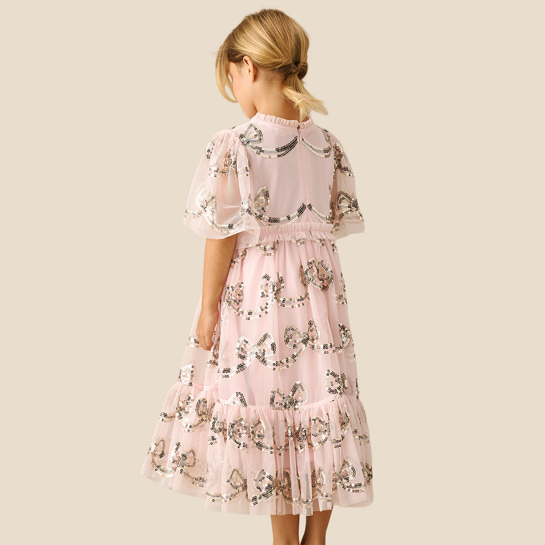 A young girl smiling and wearing a light pink tiered tulle dress with a delicate black and white floral print, set against a solid beige background.