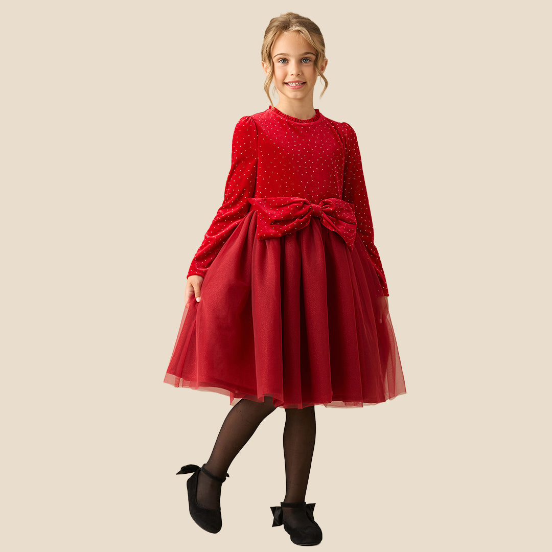 A young girl wearing a festive red dress with long sleeves, tiny gold polka dots, a matching red bow at the waist, and a tulle skirt, set against a solid beige background.
