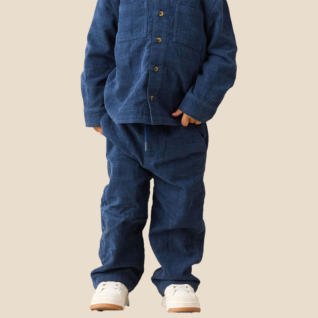 A child wearing a matching two-piece indigo blue textured pajama set with a button-up shirt and pants, paired with white sneakers, against a solid beige background.