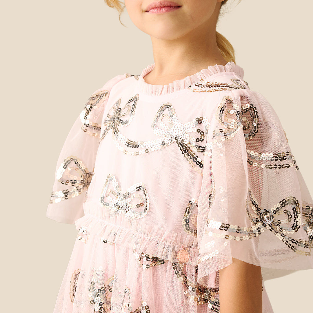 A young girl smiling and wearing a light pink tiered tulle dress with a delicate black and white floral print, set against a solid beige background.