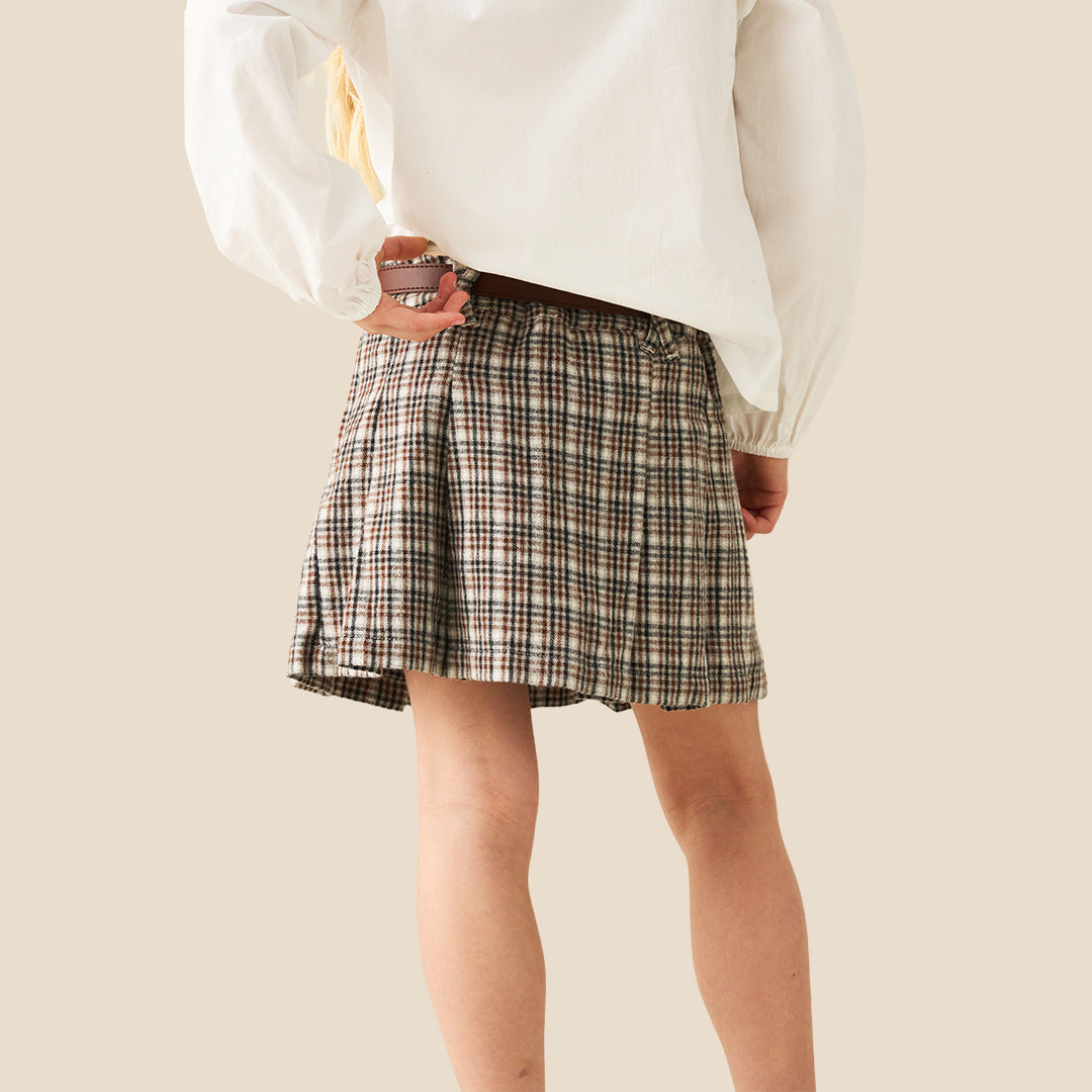 A model wearing a pleated plaid mini skirt in beige, brown, and black, paired with a white blouse and a brown belt with a gold ring, set against a solid beige background.