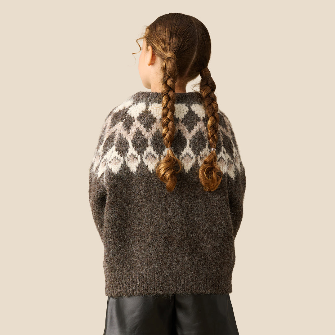 A young girl modeling a chunky knit crewneck sweater with a brown and cream argyle pattern, paired with a black faux-leather mini skirt, against a solid beige background.