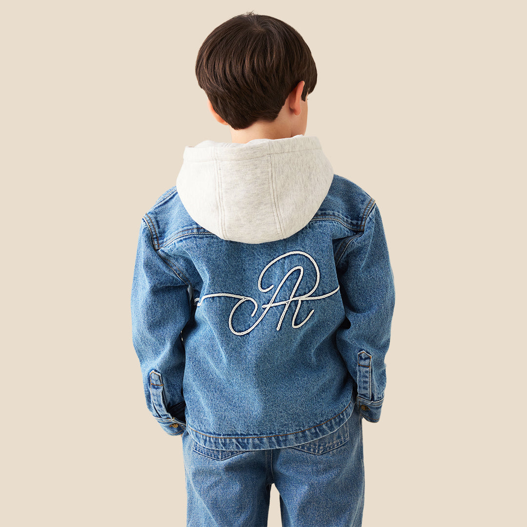 A young boy wearing a light blue denim zip-up jacket with a white hood lining and matching blue jeans, standing against a solid beige background.