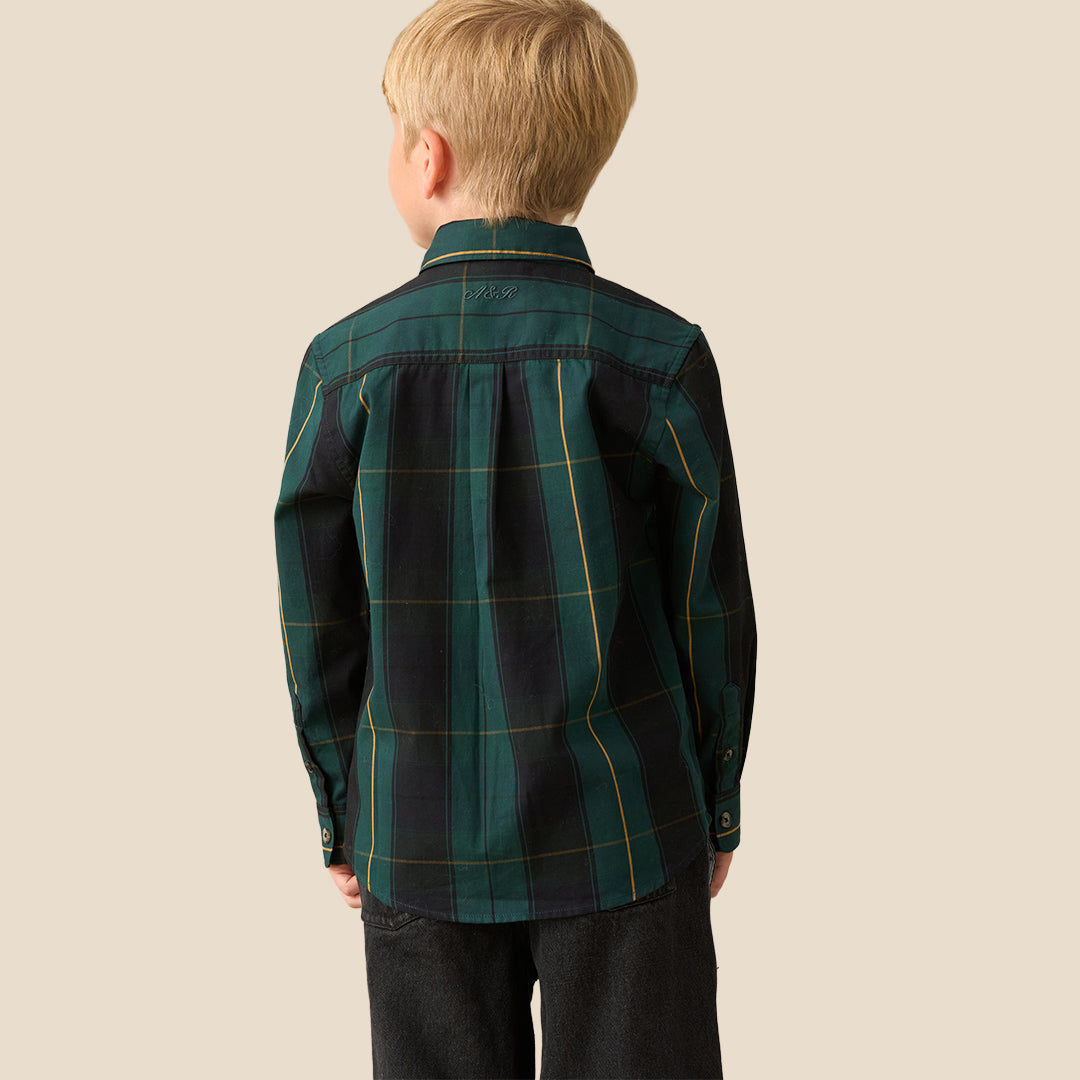 A young blond boy wearing a long-sleeve button-up shirt with a dark green and black plaid pattern, a matching dark bow tie, and dark pants, standing against a solid beige background.
