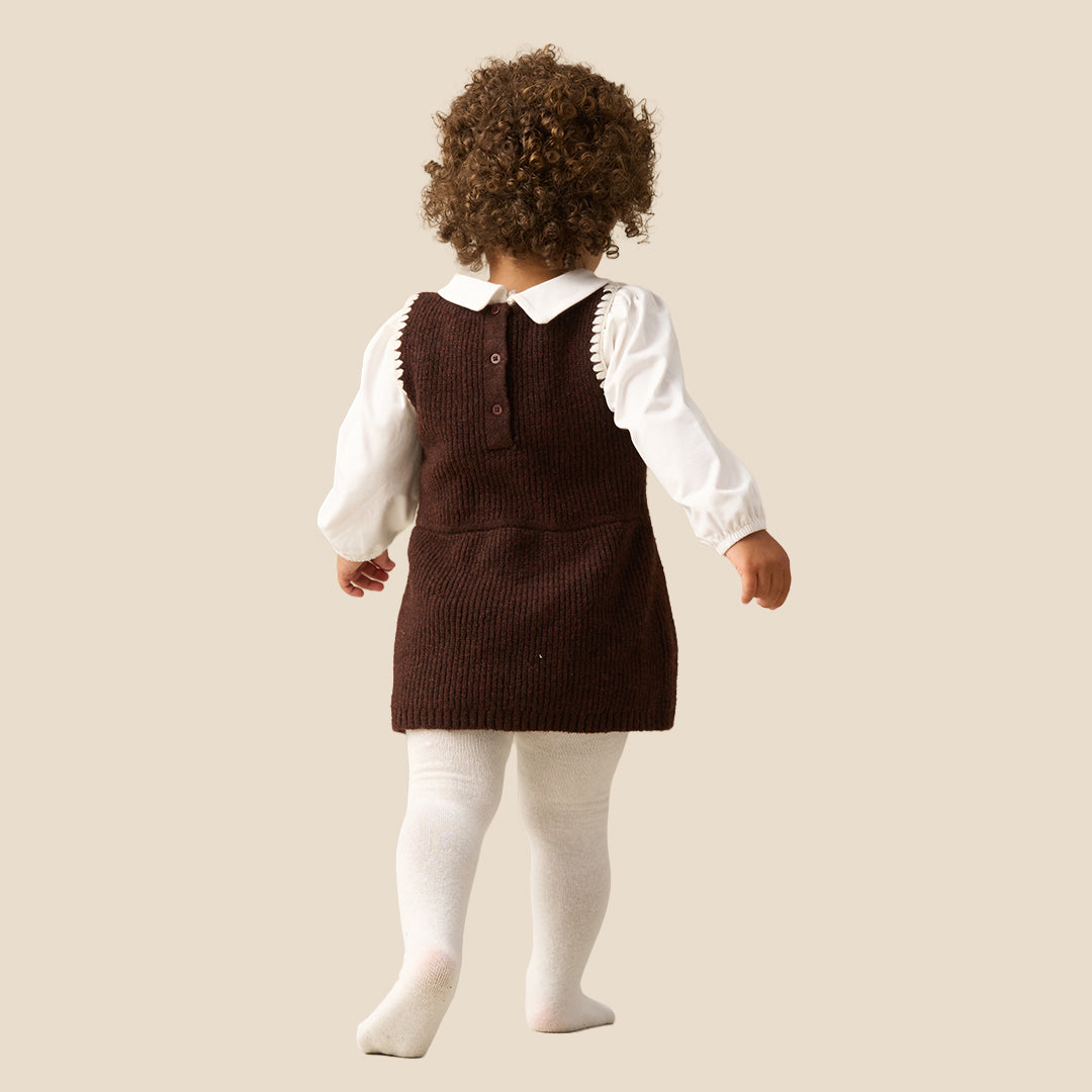 A toddler with curly brown hair wearing a dark brown corduroy pinafore dress over a white long-sleeve collared shirt and white tights, standing against a solid light beige background.