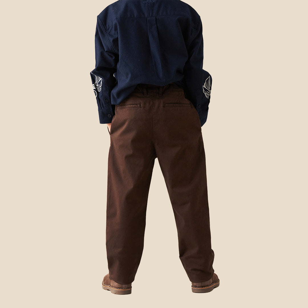 A child wearing dark brown trousers and a navy blue button-up shirt with white graphic prints, standing against a solid beige background.