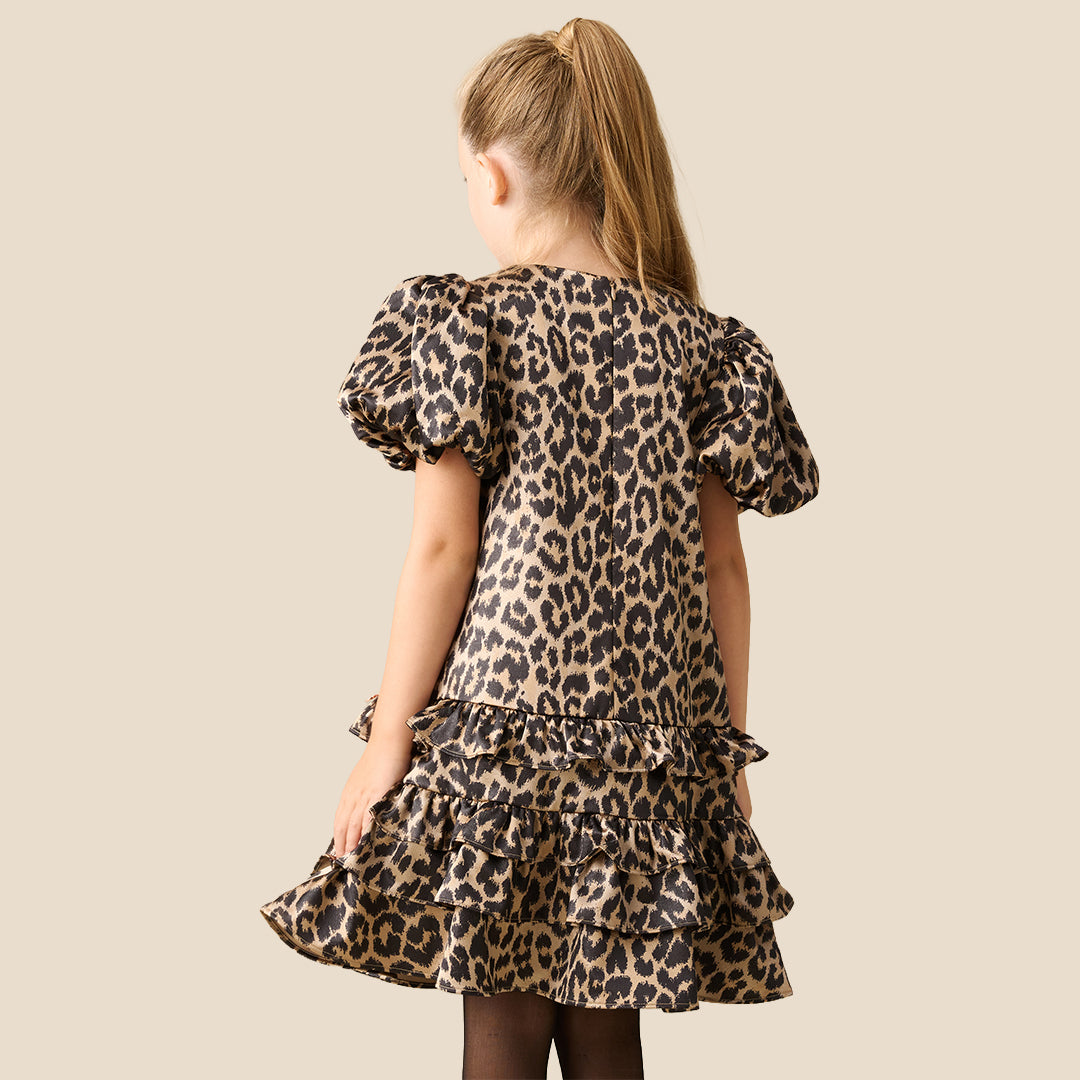A young girl with blonde hair tied back wears a leopard print dress with puff sleeves and a tiered ruffle hem, standing against a solid beige background.