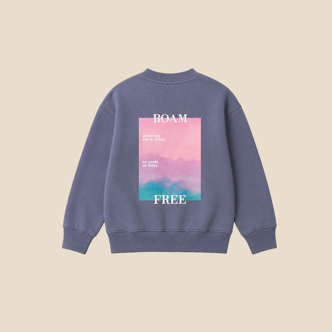 A young boy wearing a slate blue crewneck sweatshirt with a small colorful graphic on the left chest, paired with gray jeans, standing against a solid beige background.