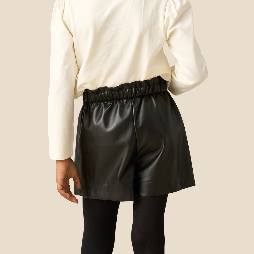 A child wearing a black faux-leather wrap mini skirt with a large black fabric bow at the waist, styled with a cream long-sleeve top and black tights against a solid beige background.