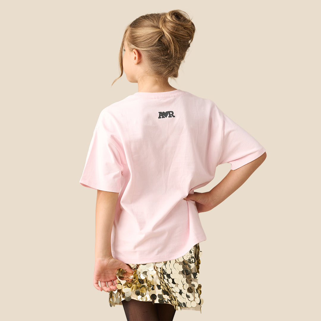 A young girl modeling a light pink graphic t-shirt with a black and pink ski design and text, paired with a gold sequined skirt, against a solid beige background.
