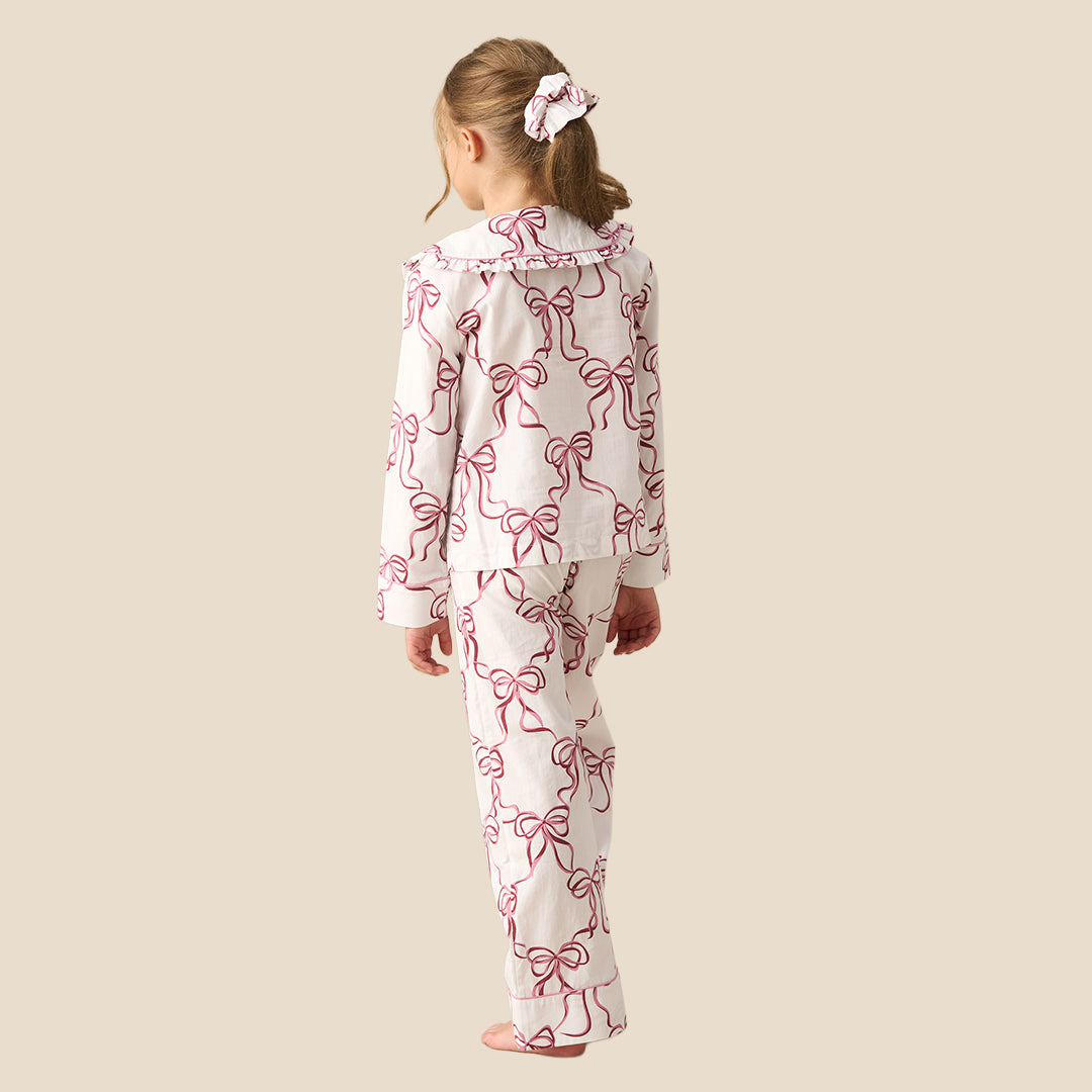 A young girl modeling a two-piece pajama set with a white base and a pink bow pattern, standing against a solid beige background.
