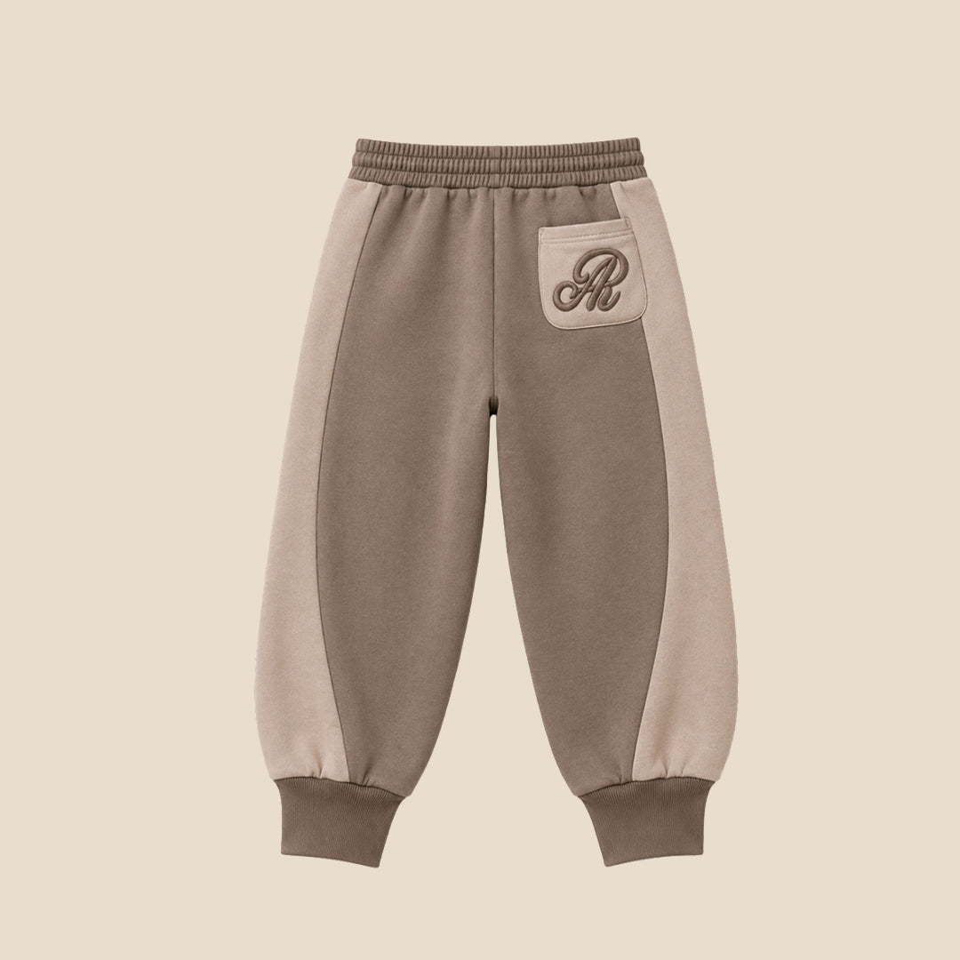 A person wearing loose taupe-brown jogger sweatpants with a white drawstring, styled with a beige and cream sweatshirt and white sneakers, against a solid beige background.