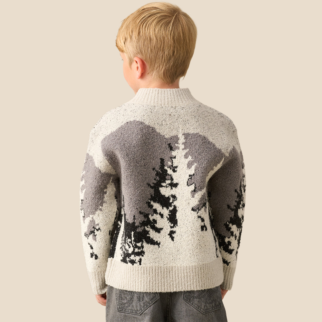 A young boy with blonde hair wearing a knit mock-neck sweater with a gray and black tree pattern, standing against a solid beige background.