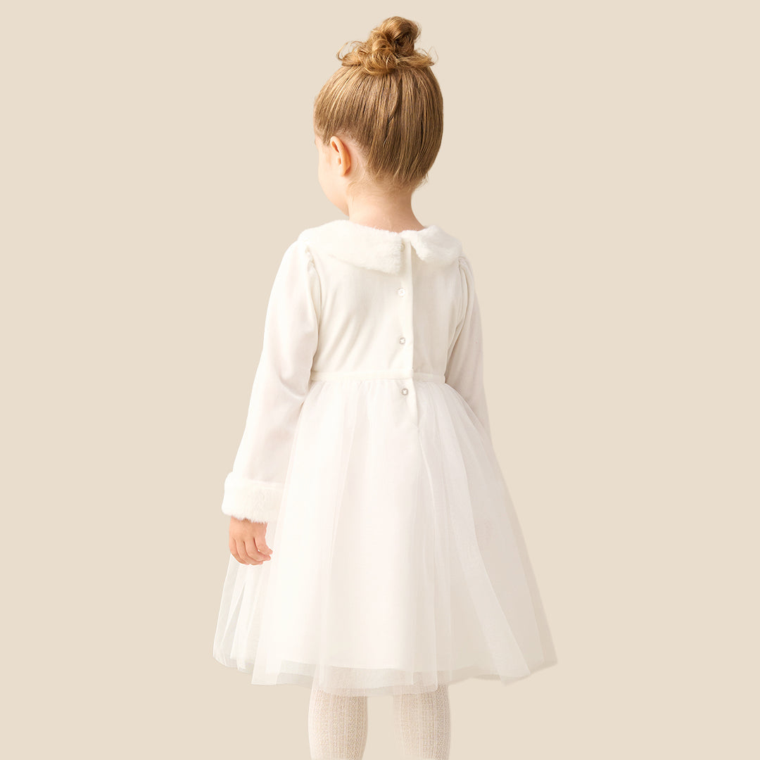 A young girl with blonde hair in a bun wearing a white long-sleeve tulle dress with faux fur trim on the collar and cuffs, standing against a solid beige background.