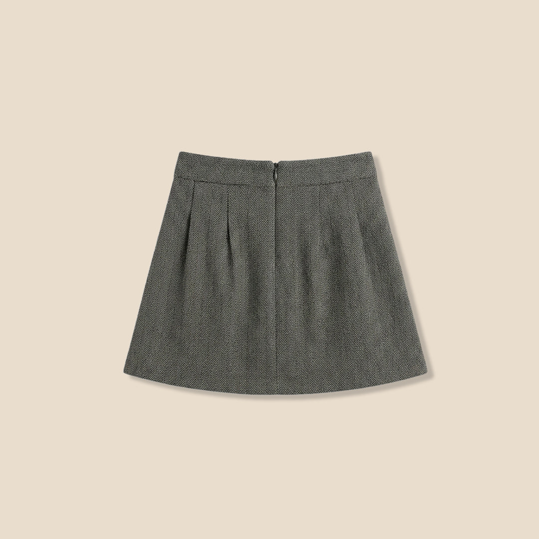 A model wearing a light pink sweatshirt with brown cursive text and a dark olive green pleated tweed mini skirt, standing against a solid beige background.