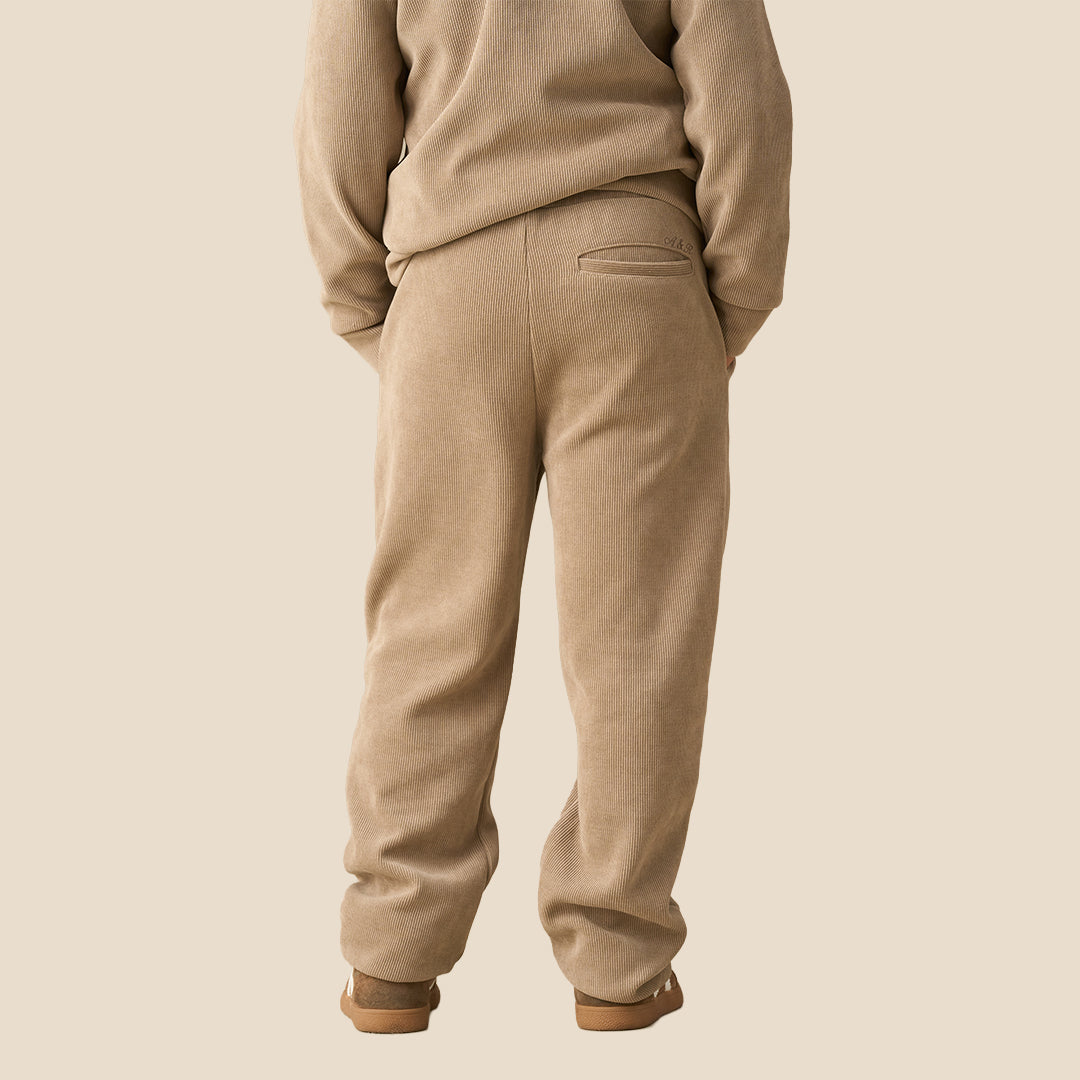 A child wearing a matching beige fleece tracksuit with a half-zip sweatshirt and relaxed-fit pants, paired with brown sneakers, against a solid light beige background.