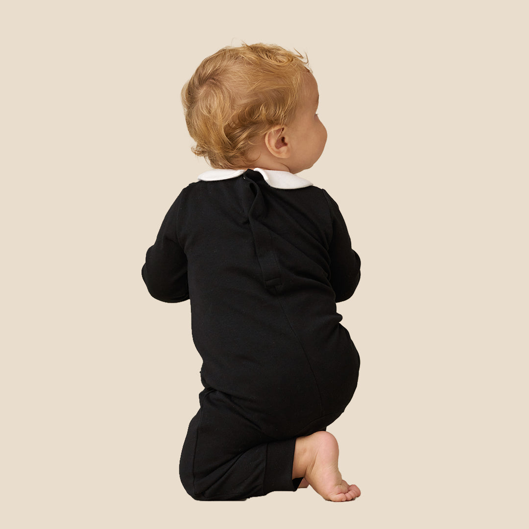 A baby wearing a black formal tuxedo-style romper with a white shirt and black bow tie, posing with arms raised against a plain beige background.