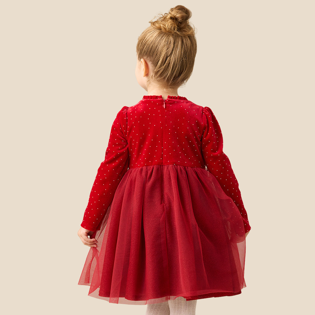 A young girl with her hair in a bun stands against a solid beige background wearing a festive red dress with a polka-dot velvet bodice, long sleeves, a large matching bow at the waist, and a tulle skirt. She is also wearing white knit tights and metallic gold shoes.