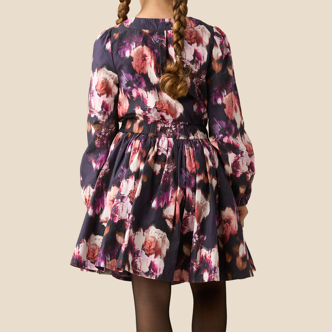 A long-sleeve floral dress with a large bow at the collar and a matching waist tie, shown on a model against a solid beige background.