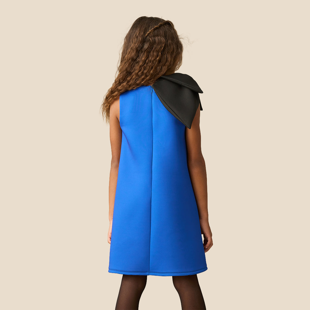 A young girl with long brown hair wearing a bright blue sleeveless dress with a large black bow on the shoulder, standing against a solid beige background.