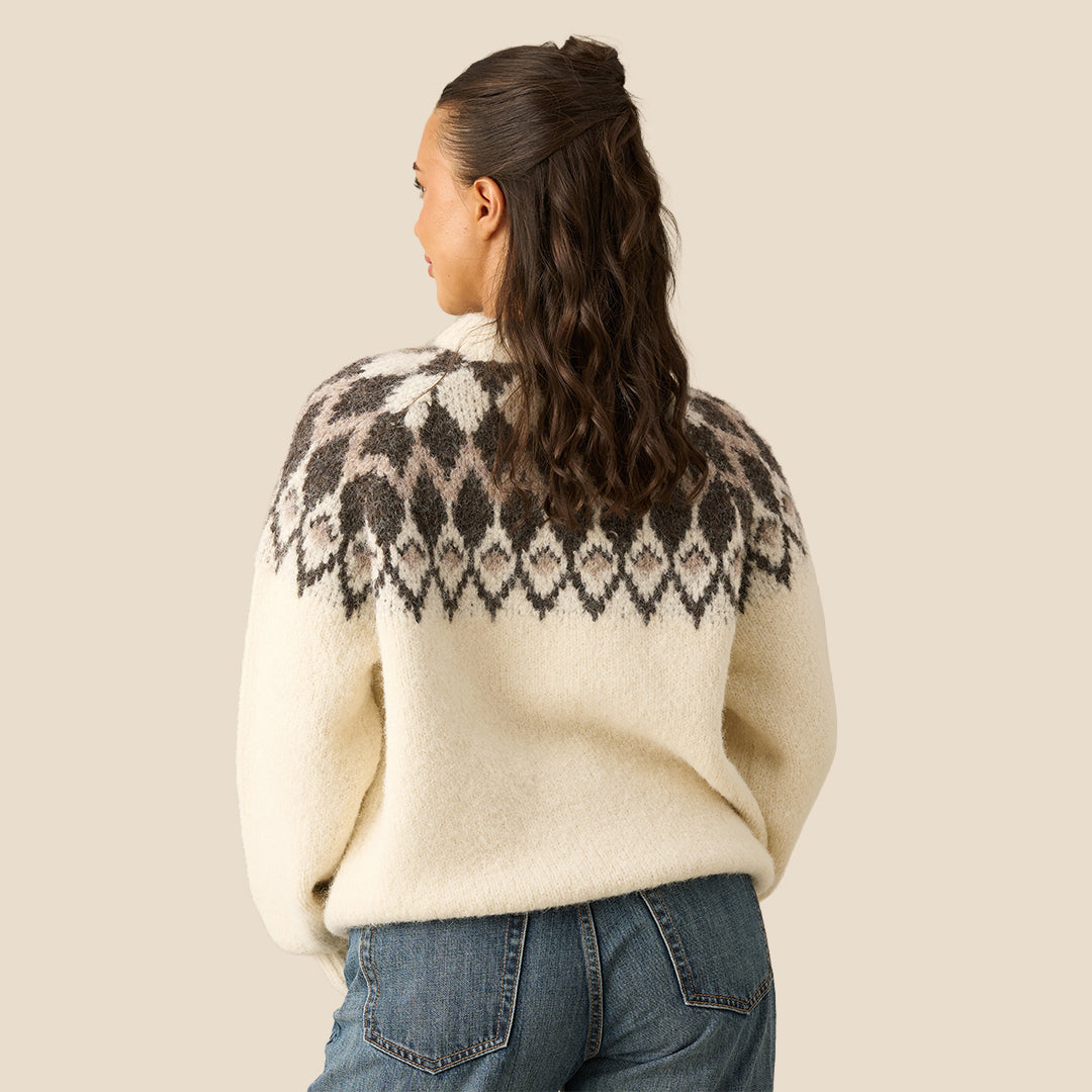 A woman wearing a cream-colored knit sweater with a dark gray and brown Fair Isle yoke pattern, paired with blue jeans, against a solid beige background.
