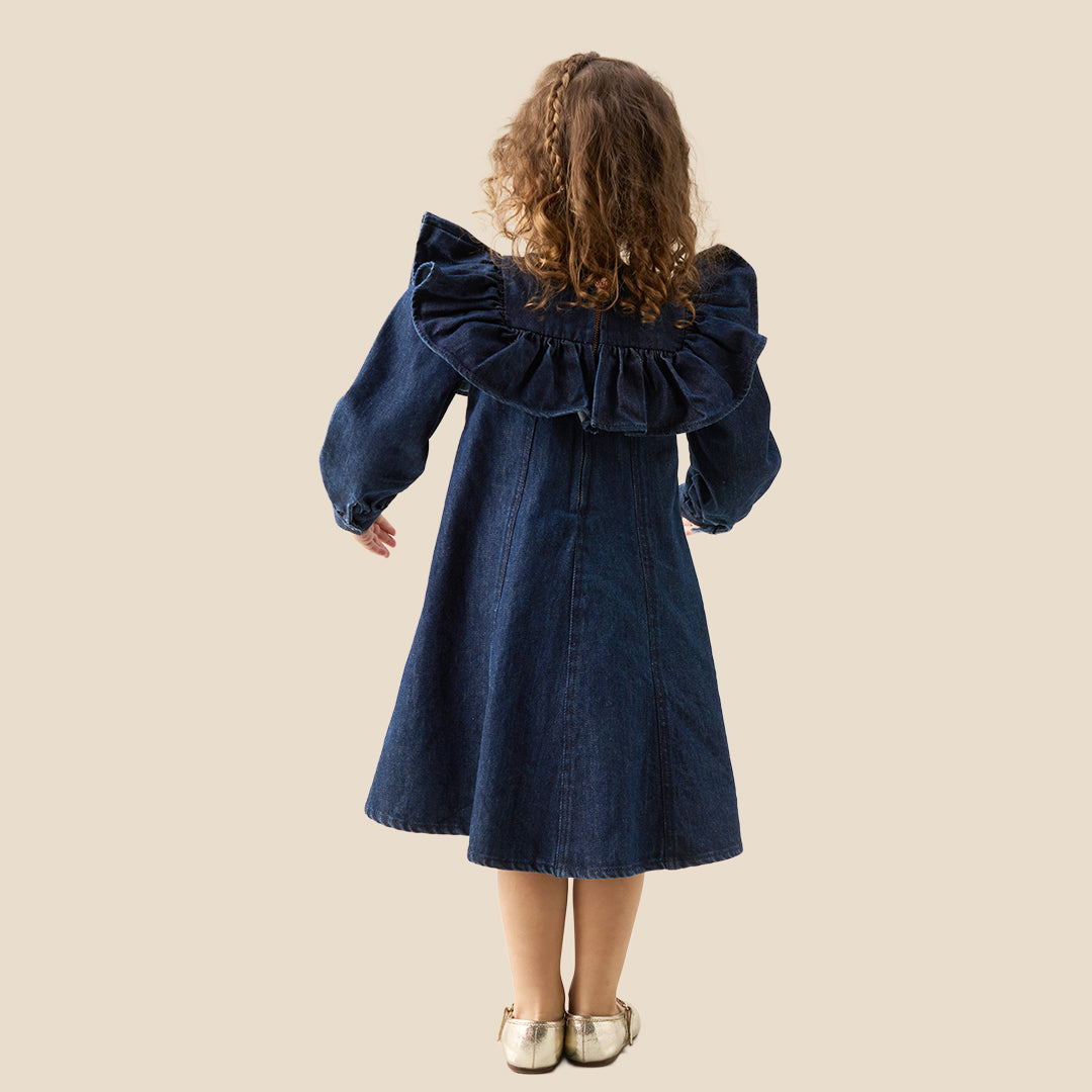 A young child wearing a dark blue denim dress with dramatic ruffled shoulders, standing against a solid beige background.