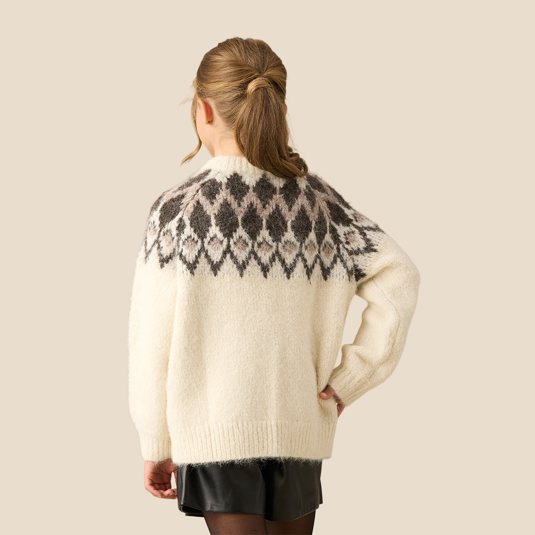 A young girl smiling while wearing a cream-colored knit sweater with a dark brown and gray geometric pattern across the chest, paired with a black skirt, against a solid beige background.