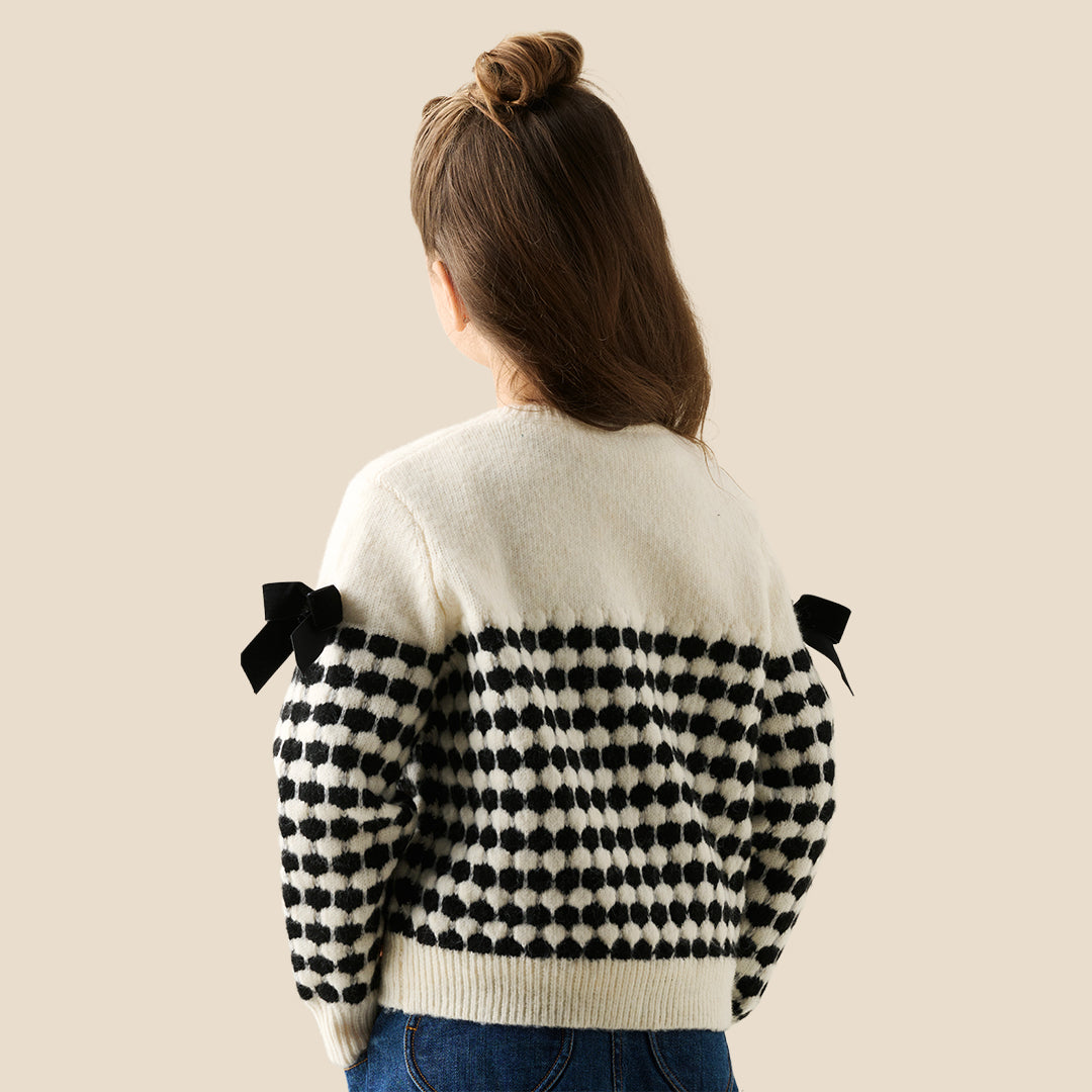 A young girl with her hair in a bun wears a cream knit cardigan with black polka-dot patterned sleeves and black ribbon bows on the shoulders, paired with a dark blue ruffled skirt, against a solid beige background.
