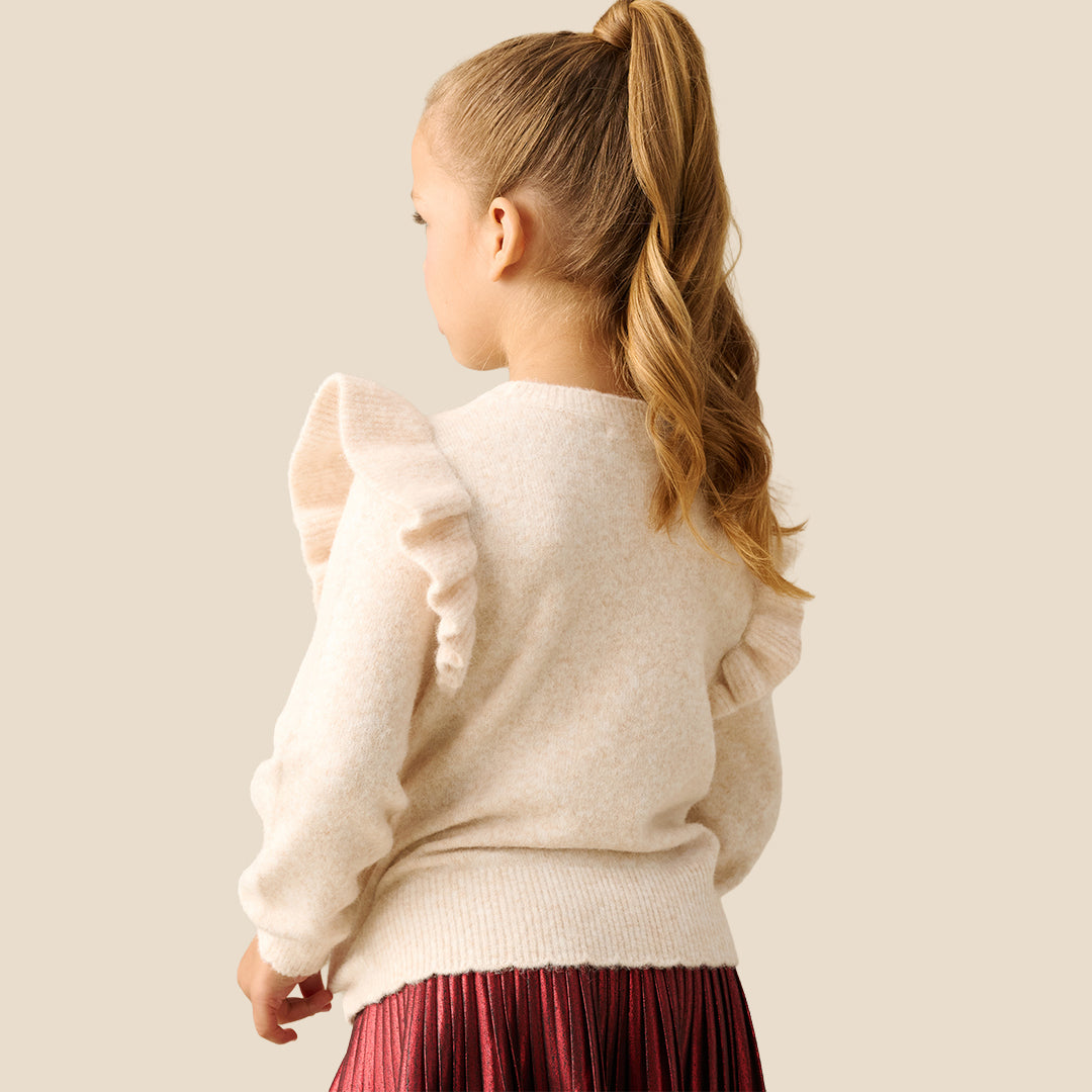 A young girl with blonde hair in a ponytail models a cream-colored knit sweater featuring a large embroidered reindeer on the front with colorful antlers and a red bow. She is wearing a shiny red skirt against a solid light beige background.