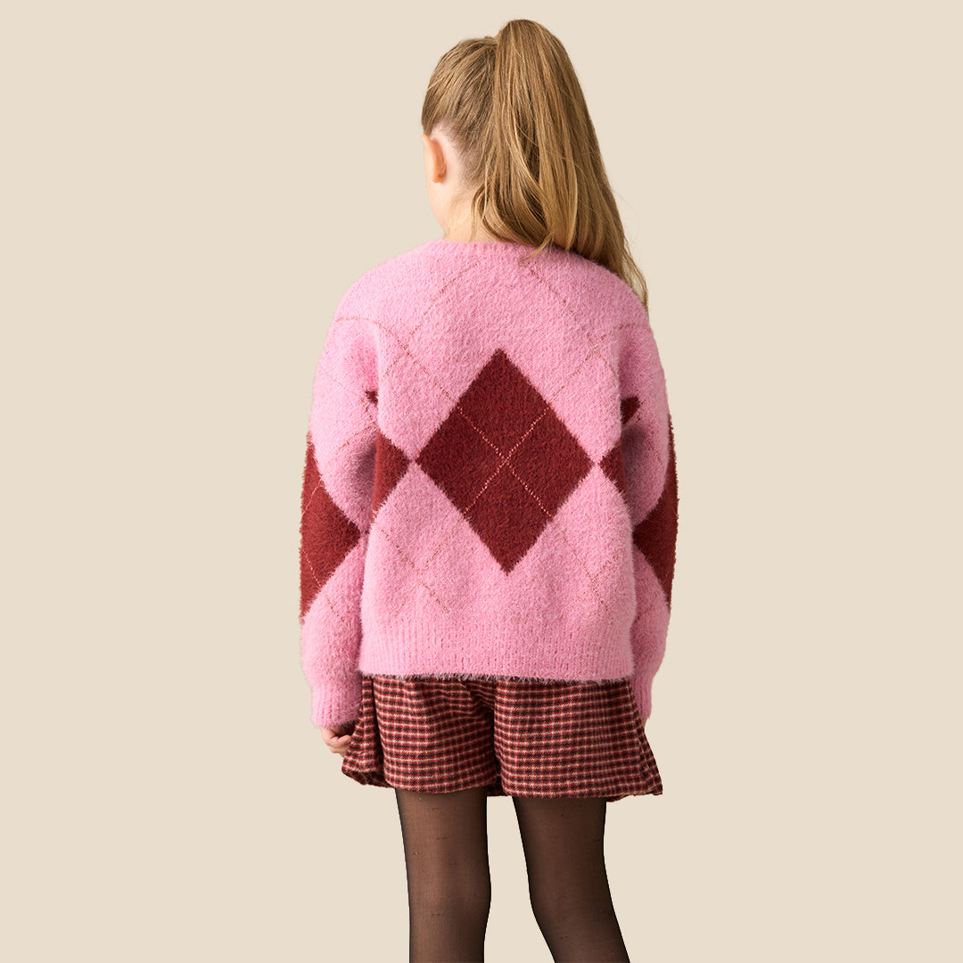 A young girl modeling a pink and dark red argyle sweater with small bow details, paired with a red and black gingham pleated skirt, against a solid beige background.