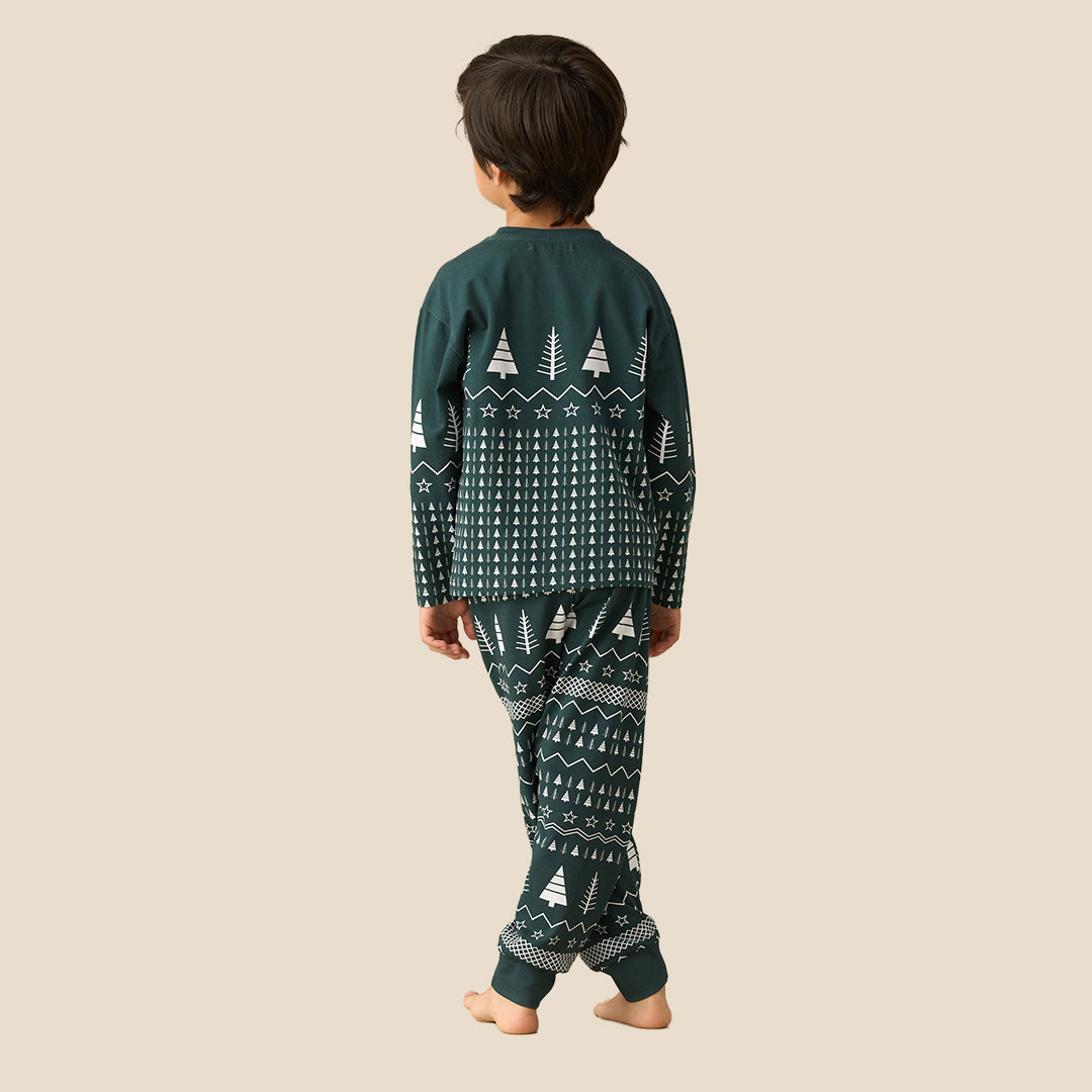 A young boy stands barefoot against a solid beige background, wearing a dark green long-sleeve pajama set with a white Nordic tree pattern.