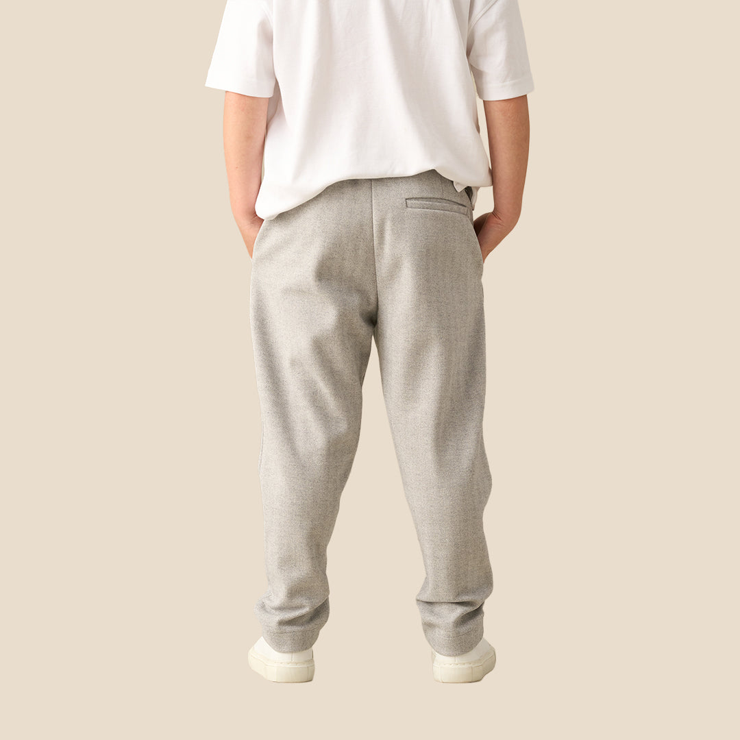 A child wearing light gray heathered sweatpants with a white t-shirt and white sneakers, standing against a solid beige background.