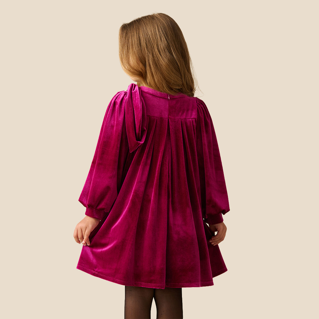 A young girl with blonde hair wearing a long-sleeved magenta velvet dress with a gathered bow detail on the chest, standing against a solid beige background.