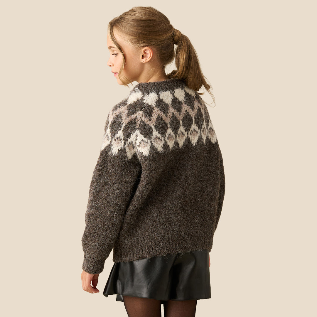 A young girl modeling a chunky knit crewneck sweater with a brown and cream argyle pattern, paired with a black faux-leather mini skirt, against a solid beige background.