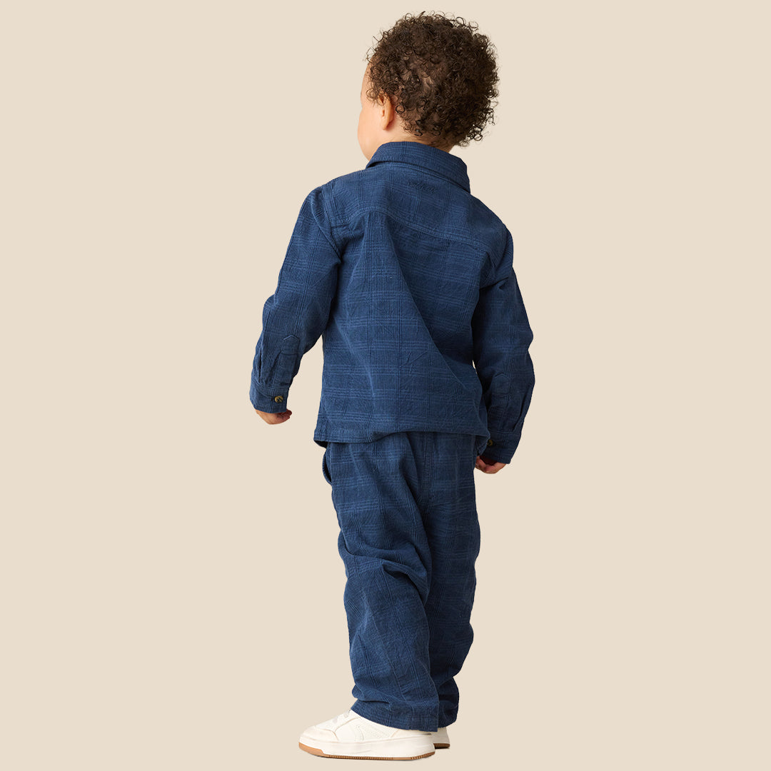 A toddler standing against a plain beige studio background, wearing a matching dark blue denim-like two-piece outfit with a collared long-sleeve top and pants, plus white sneakers.