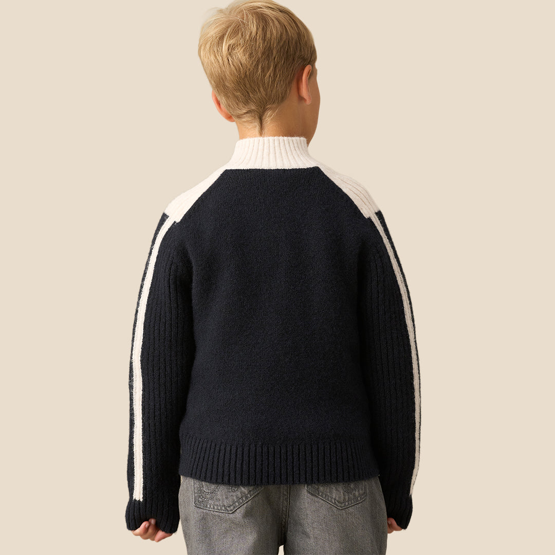 A young boy wearing a navy blue and cream knit quarter-zip sweater with white stripe details on the sleeves, paired with gray pants, against a solid beige background.