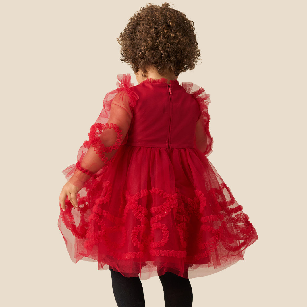 A toddler with curly brown hair wearing a red tulle dress with sheer long sleeves and red floral applique details, standing against a solid beige background.