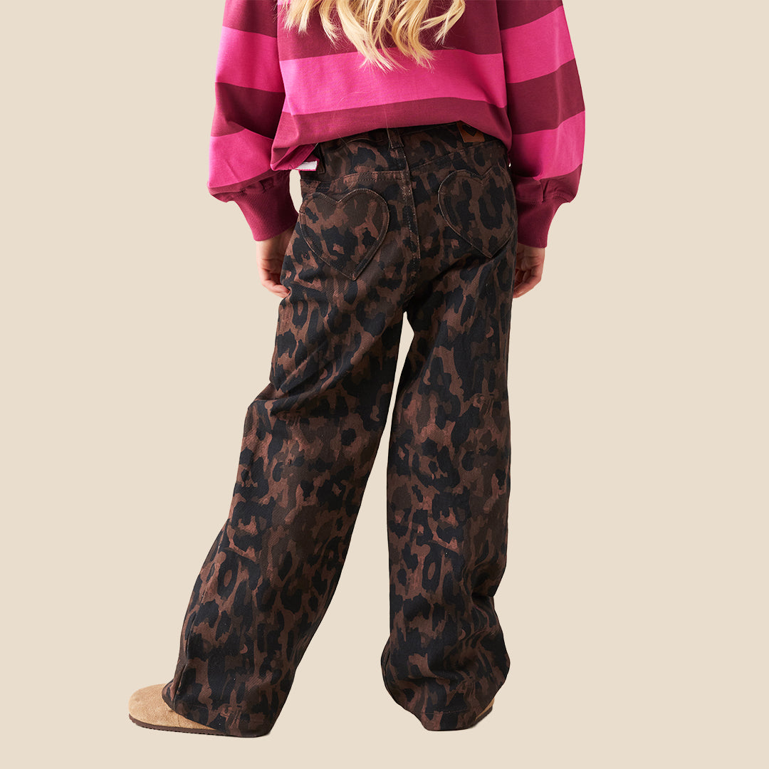 A person wearing loose, wide-leg brown and black leopard-print pants, paired with a bright pink striped long-sleeve top and tan slip-on shoes, against a plain beige background.