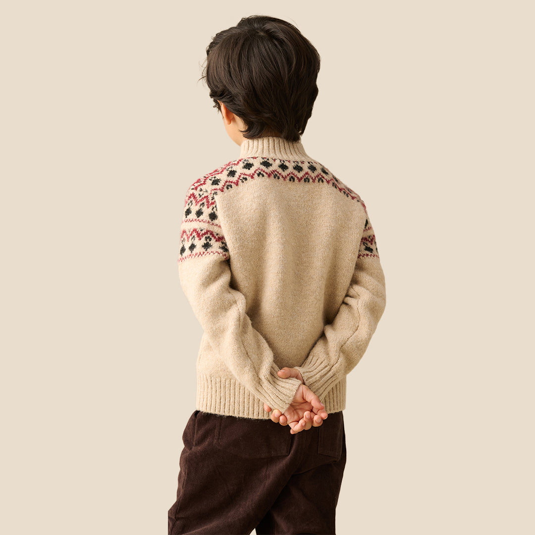 A young boy wearing a beige knit half-zip sweater with red and black Fair Isle patterns on the shoulders and sleeves, set against a solid light beige background.