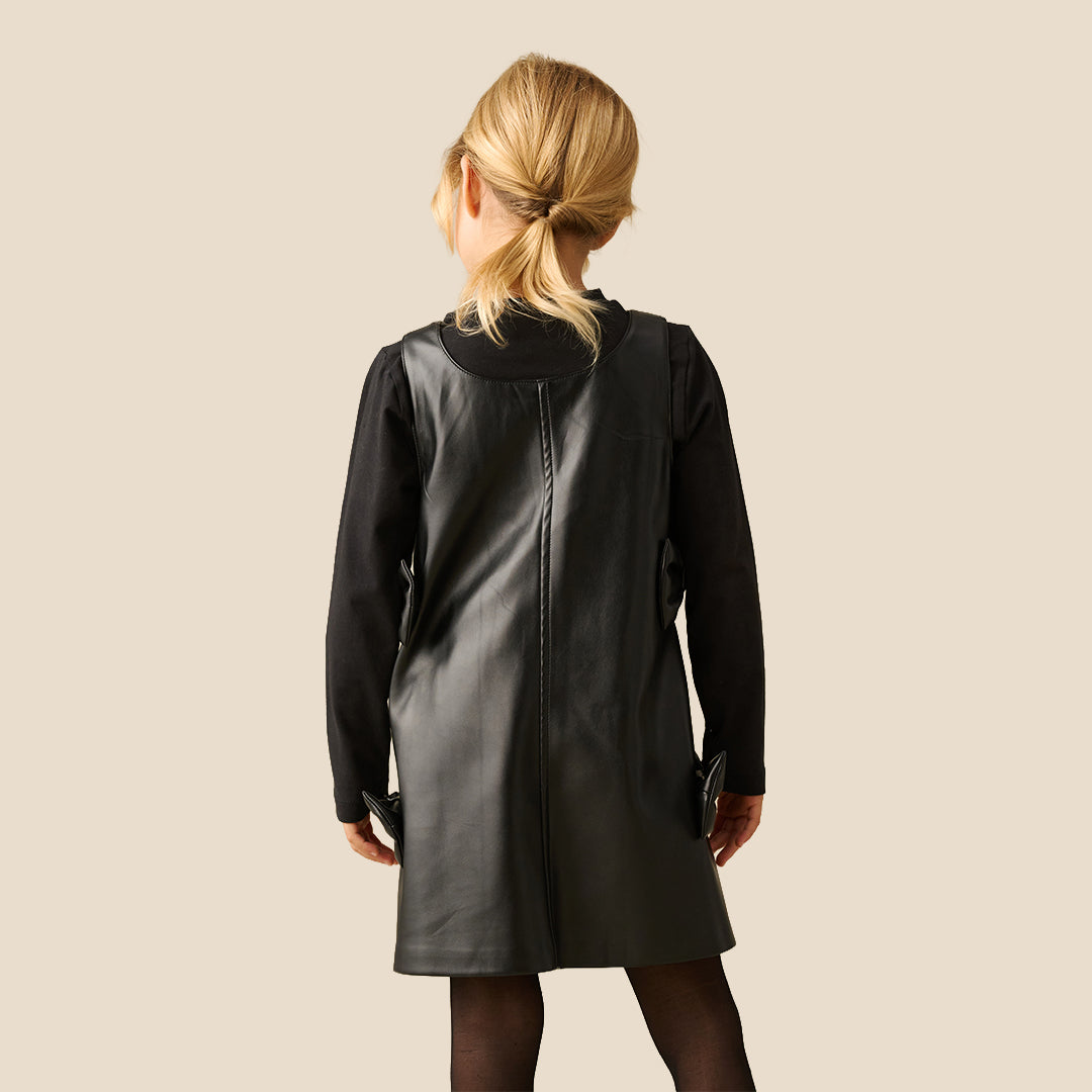 A young girl with blonde hair tied back is wearing a black faux-leather dress with long sleeves, a mock neck, and decorative bows on the sides, paired with black tights, against a solid beige background.