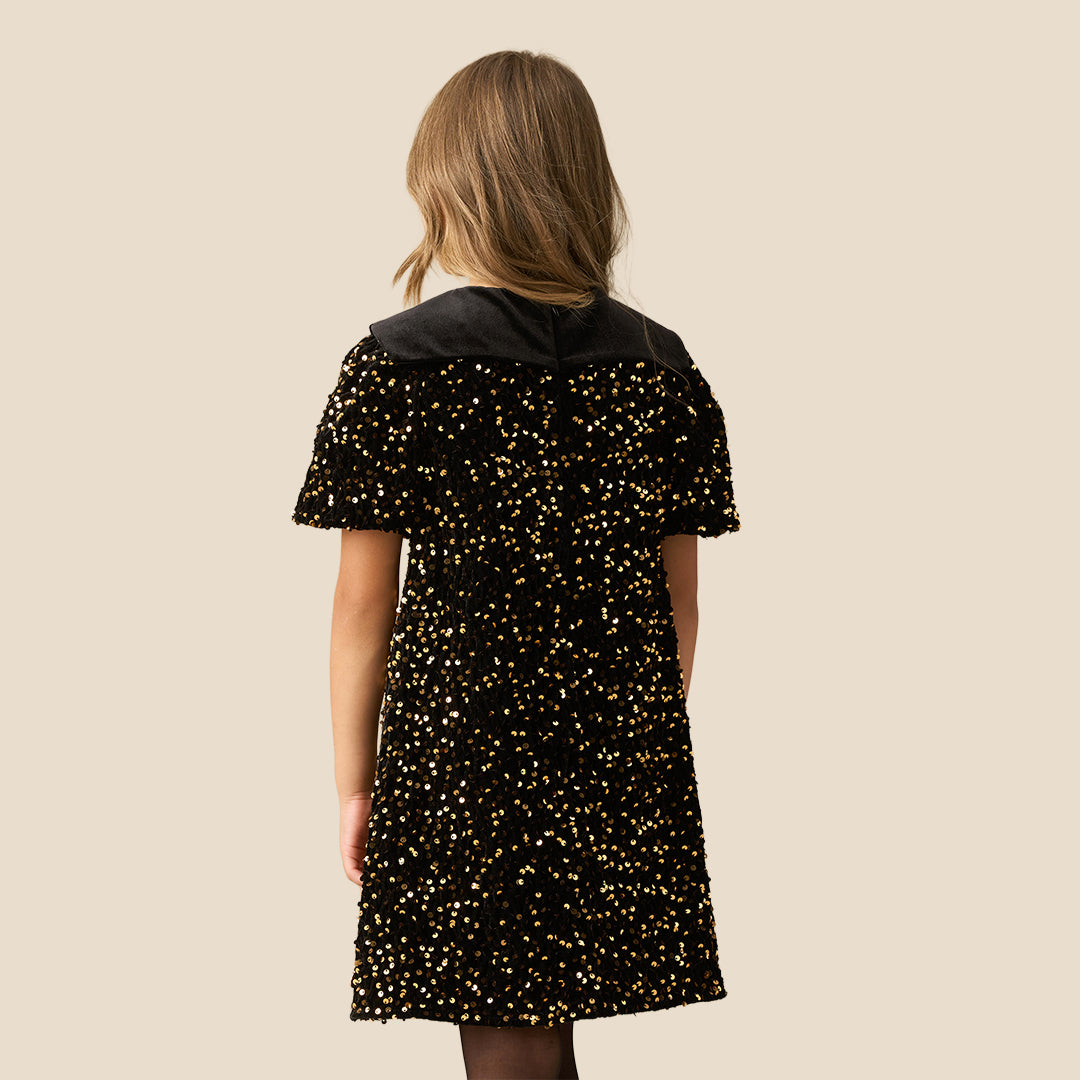 A young girl with blonde hair wearing a black short-sleeve dress with gold sequins and a black ruffled Peter Pan collar, standing against a solid beige background.