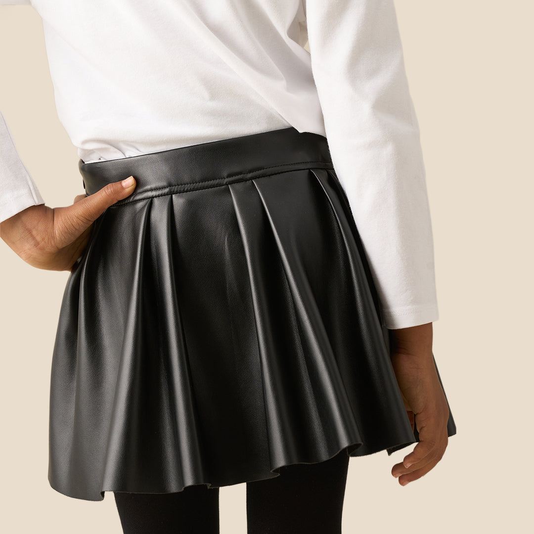 A child model wearing a black faux-leather pleated mini skirt with a matching black belt and rose gold button, paired with a white embellished top and black tights, against a solid beige background.