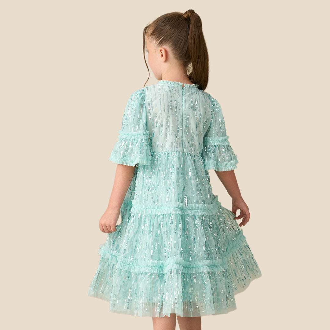 A young girl with a long brown ponytail wearing a light mint green sequined tiered dress, standing against a solid beige background.