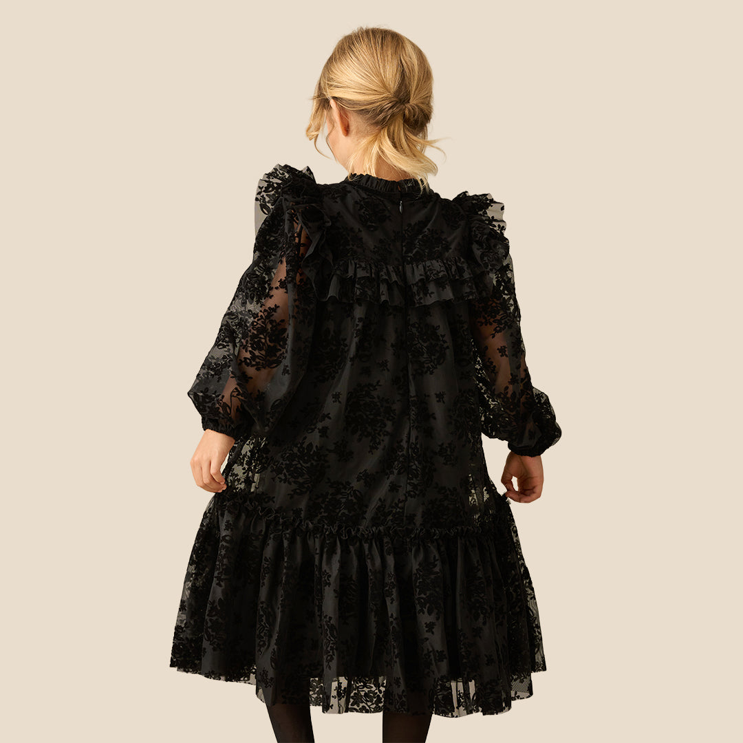 A young girl with blonde hair smiling while wearing a black lace long-sleeve dress with ruffled details, photographed against a solid light beige background.