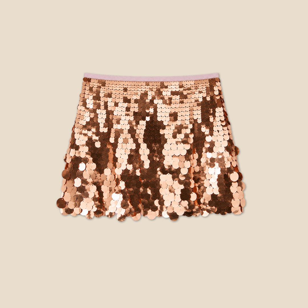 A child wearing a white graphic T-shirt and a short skirt covered in large, reversible sequins that shift between rose gold and bronze, photographed against a plain beige studio background.