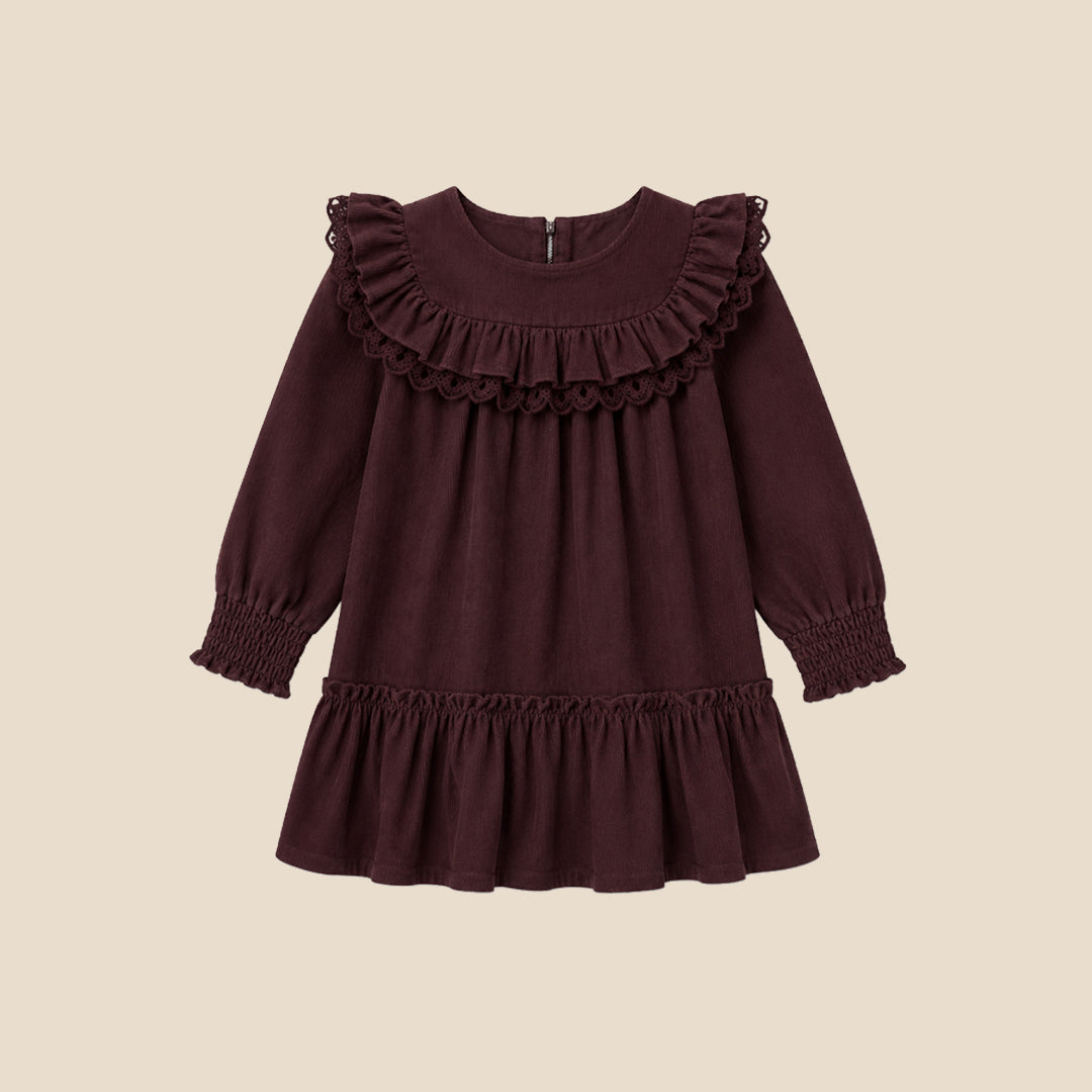 A young girl with light brown hair in pigtails stands against a solid beige background wearing a long-sleeved dark plum-brown dress with ruffles, white tights, and cream-colored shoes.