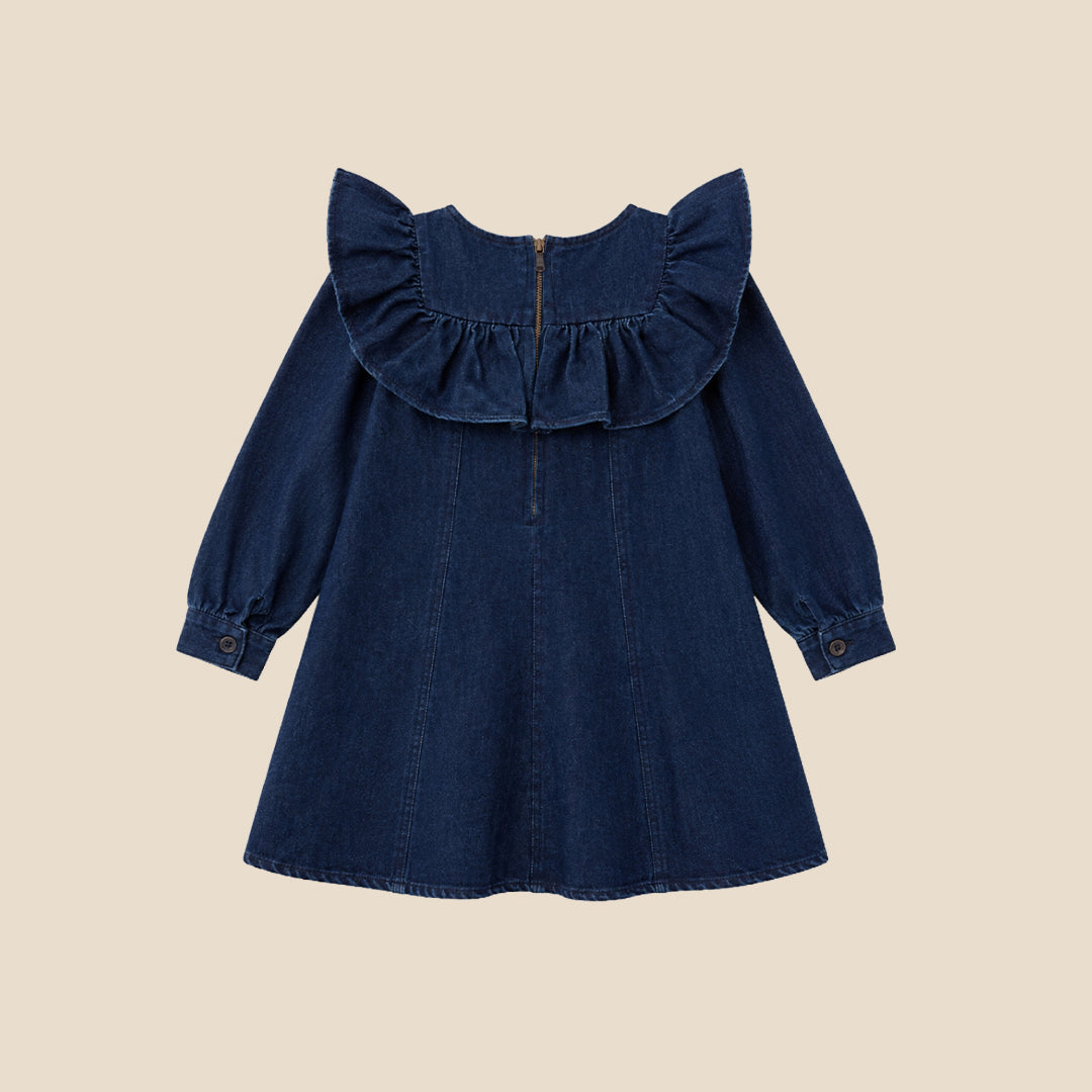 A young child wearing a dark blue denim dress with dramatic ruffled shoulders, standing against a solid beige background.